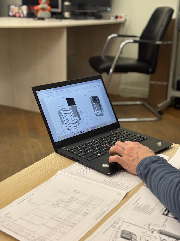 An engineer reviews detailed blueprints on a laptop at an office desk.